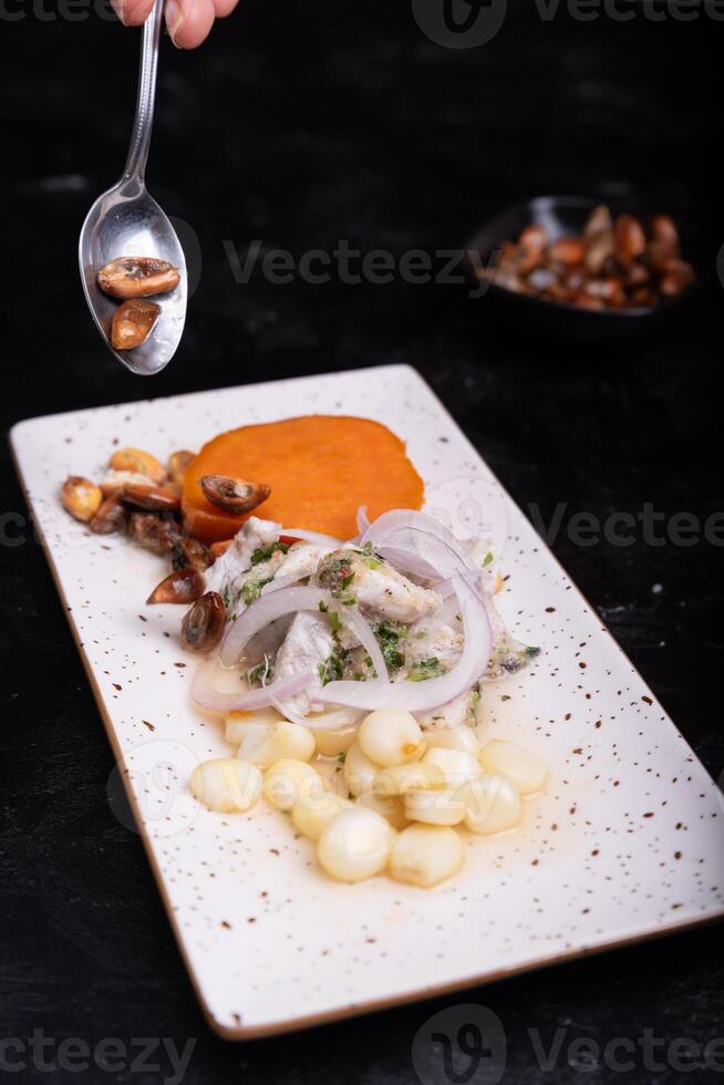 Spoon adding roasted corn over a plate of Peruvian ceviche on a dark background. photo