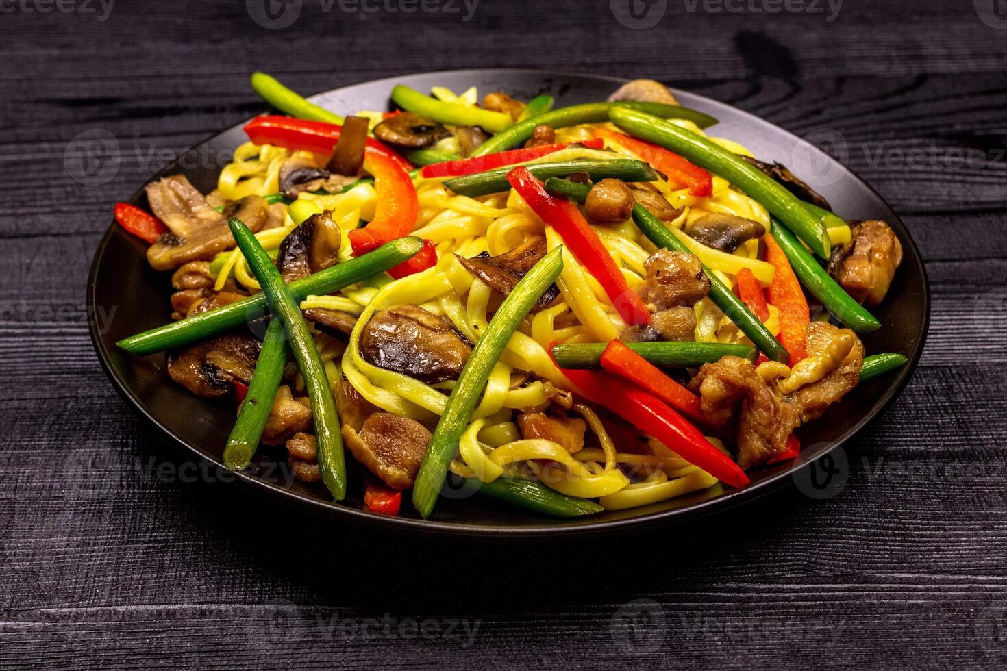 Appetizing stir fried noodles with garlic sprouts, mushrooms, red pepper and chicken meat on a black rustic background of a wooden table. A popular Asian dish photo