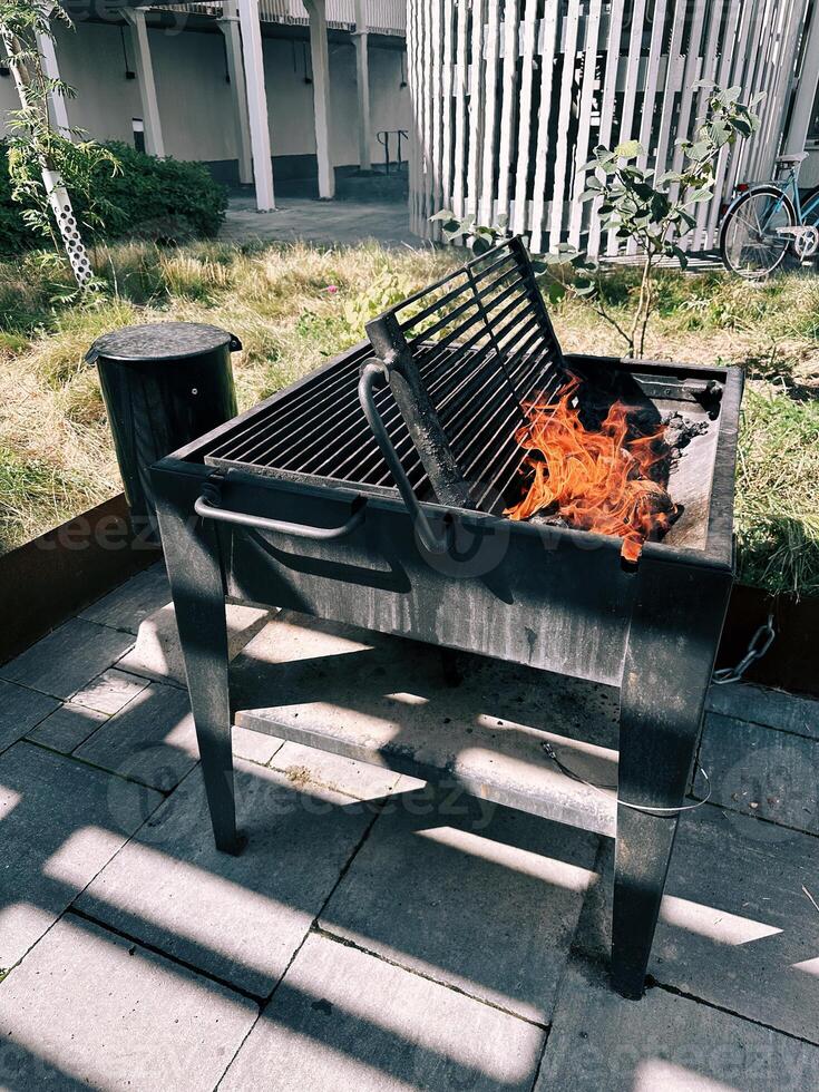 Firewood burns in an open grill in the yard against the backdrop of a modern structure, a trash can, and green grass. photo