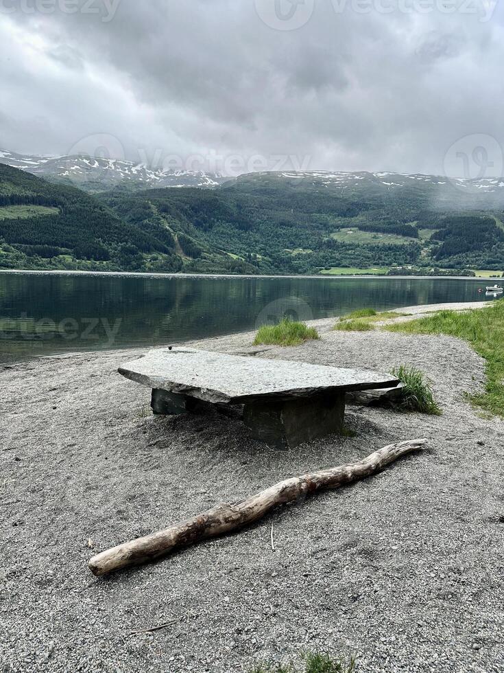 A table is placed on a pebbly shore by a lake, surrounded by a natural landscape with mountains, grass, and sky reflecting in the water. Norway photo