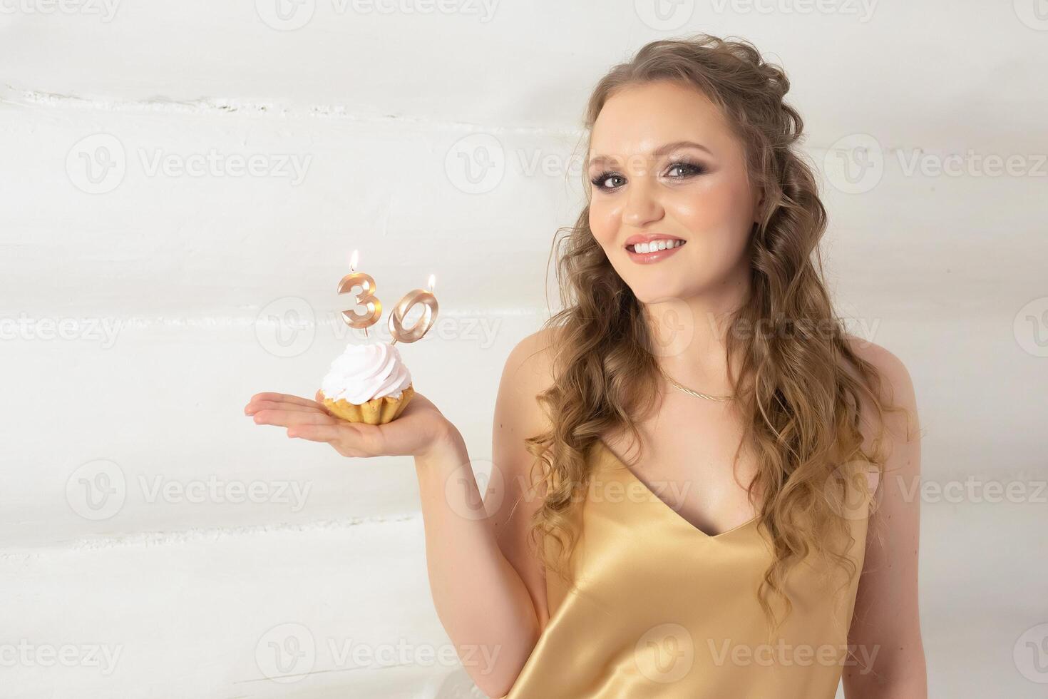 a beautiful girl with long blond curly hair in a gold dress holds a small cake with candles with the number 30. girl on her birthday 30 years old photo