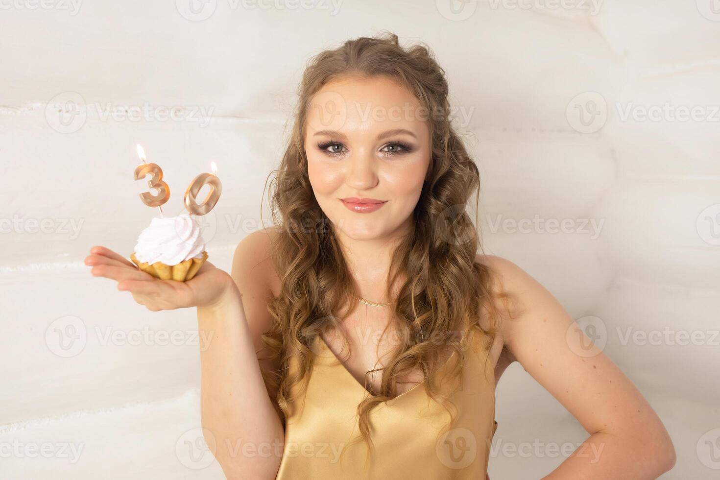 a beautiful girl with long blond curly hair in a gold dress holds a small cake with candles with the number 30. girl on her birthday 30 years old photo