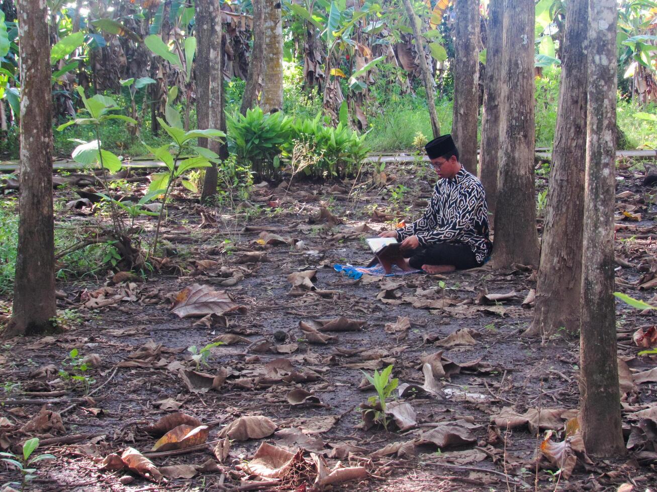 An Asian Muslim man is sitting and reading the Quran. The peace in the middle of forest makes it an energetic atmosphere of faith, traditional vibes photo