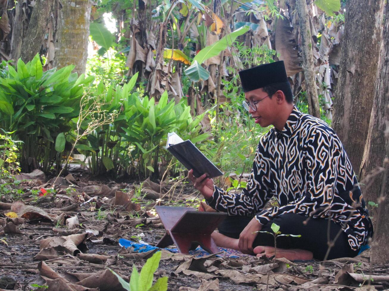 An Asian Muslim man is sitting and reading the Quran. The peace in the middle of forest makes it an energetic atmosphere of faith, traditional vibes photo