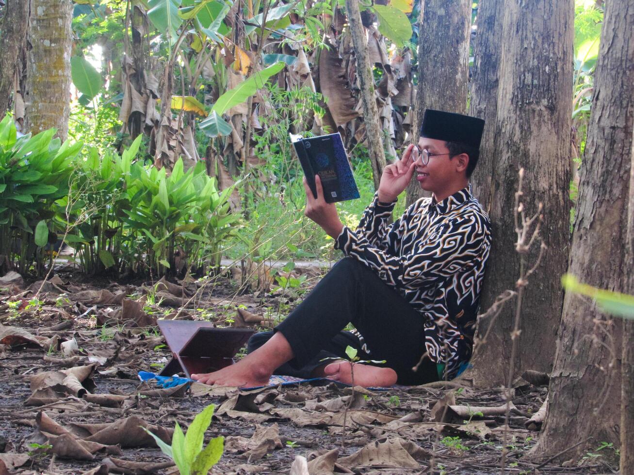 An Asian Muslim man is sitting and reading the Quran. The peace in the middle of forest makes it an energetic atmosphere of faith, traditional vibes photo