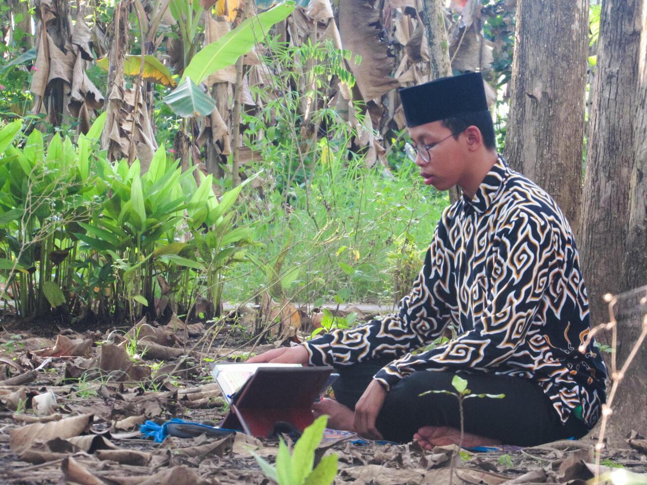 An Asian Muslim man is sitting and reading the Quran. The peace in the middle of forest makes it an energetic atmosphere of faith, traditional vibes photo