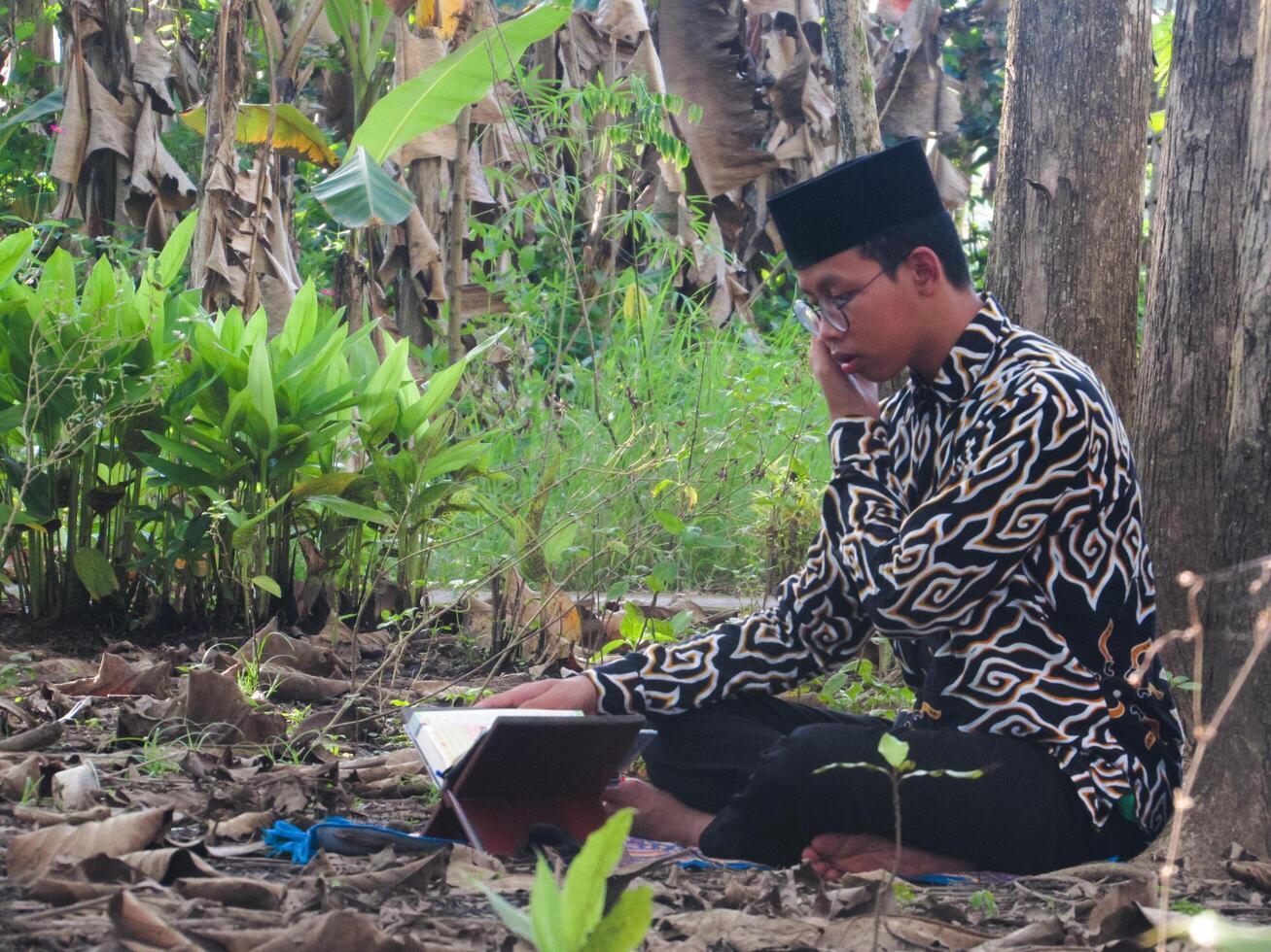An Asian Muslim man is sitting and reading the Quran. The peace in the middle of forest makes it an energetic atmosphere of faith, traditional vibes photo