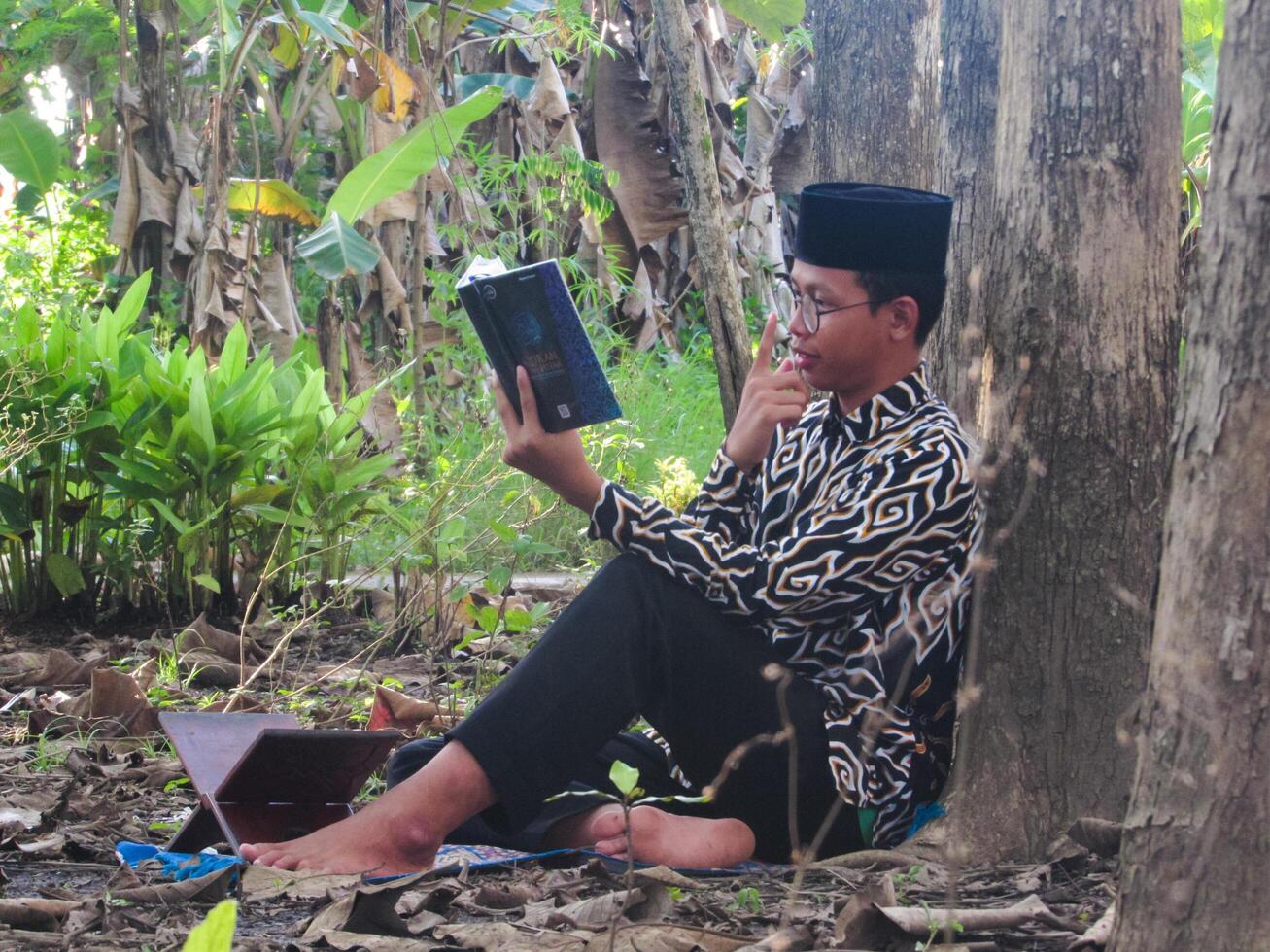 An Asian Muslim man is sitting and reading the Quran. The peace in the middle of forest makes it an energetic atmosphere of faith, traditional vibes photo