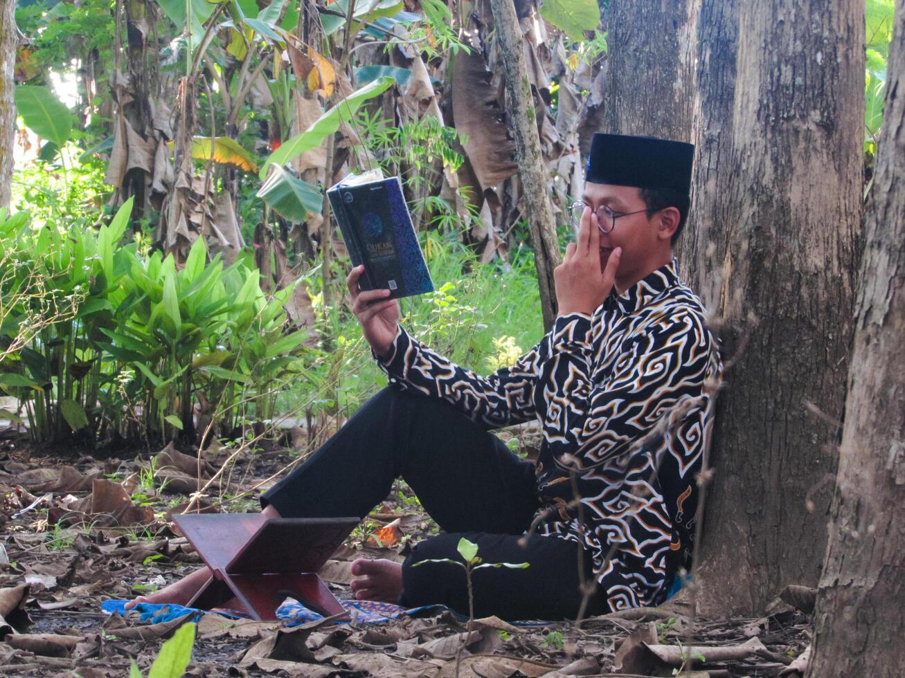 An Asian Muslim man is sitting and reading the Quran. The peace in the middle of forest makes it an energetic atmosphere of faith, traditional vibes photo