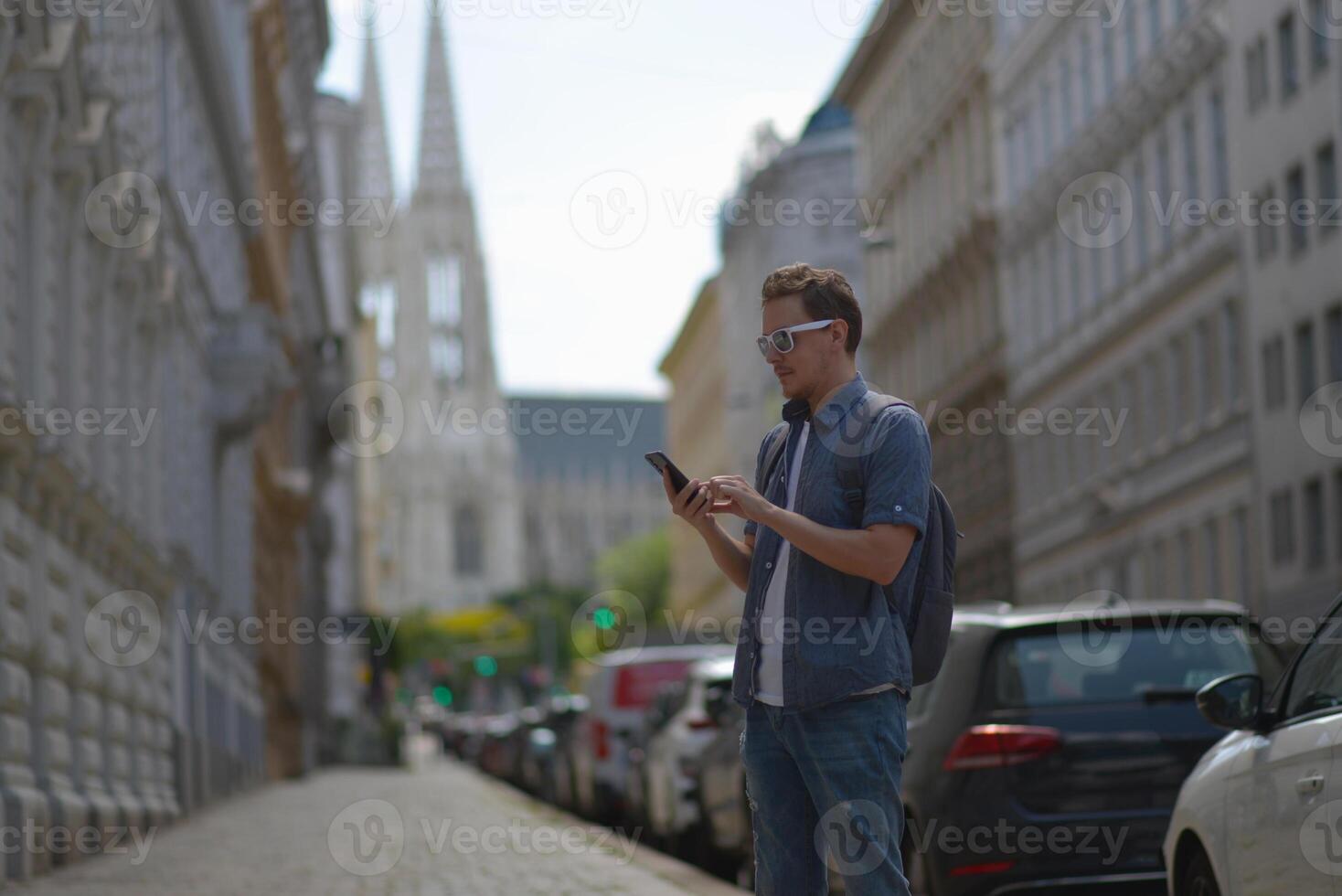 A man using smartphone wearing white sunglasses and gray backpack on the street in Vienna on a hot sunny summer day. Tourism and sightseeing concept photo
