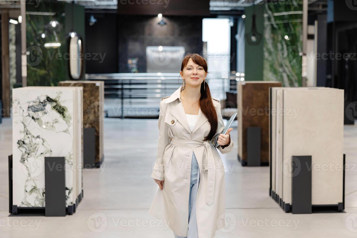 A woman in a white trench coat walks through a showroom displaying tile samples. She is holding a folder in her left hand and smiling at the camera photo