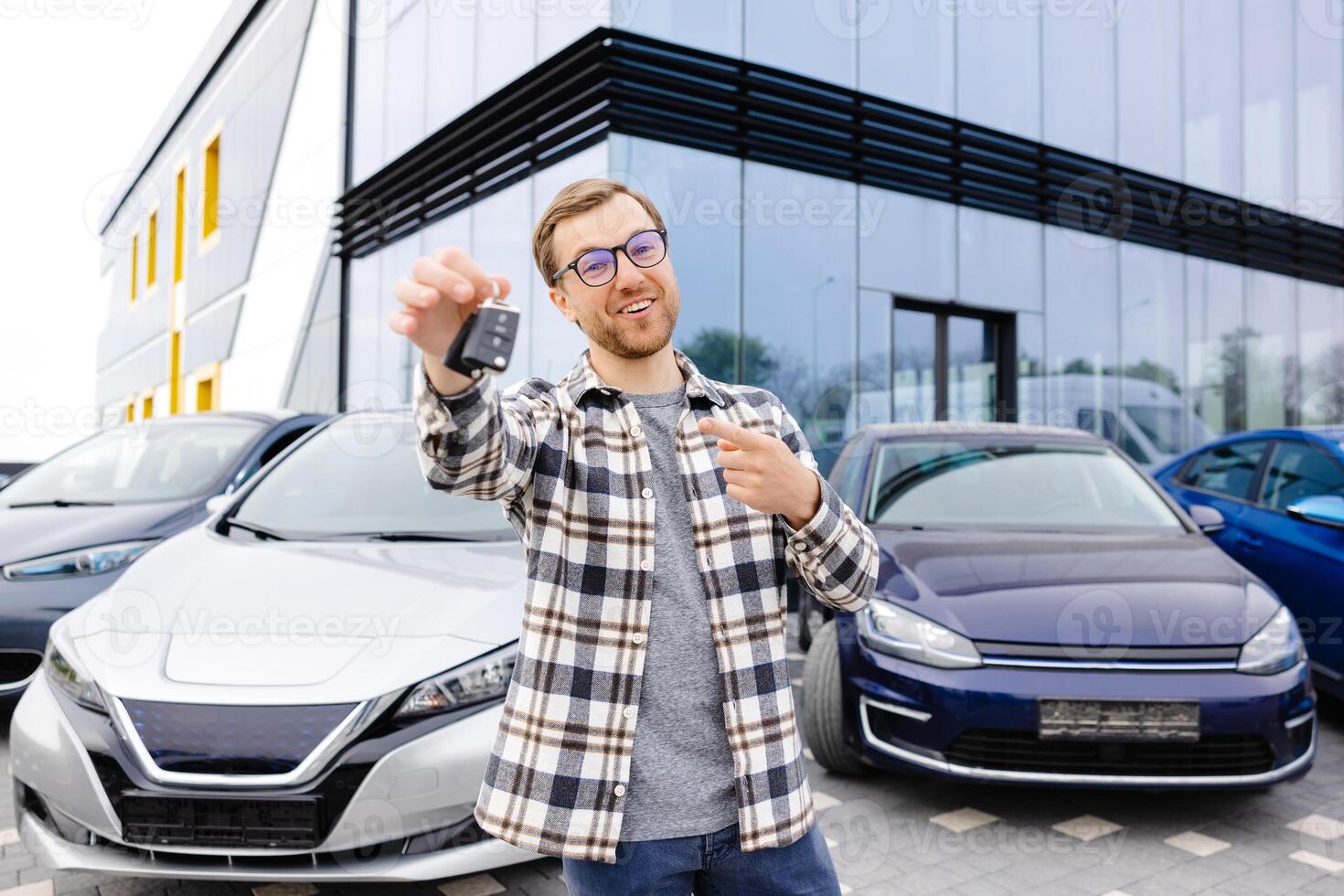Emotional guy holding key in hand and smiling at camera. Man showing key from his new electric car. Young man buying auto at dealership salon photo