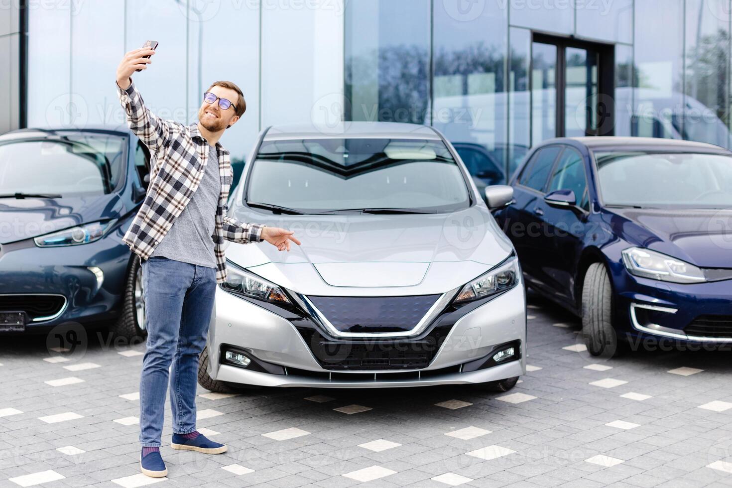 Happy man taking selfie through smart phone after purchasing new car. The man showing the purchased car via link over the phone photo