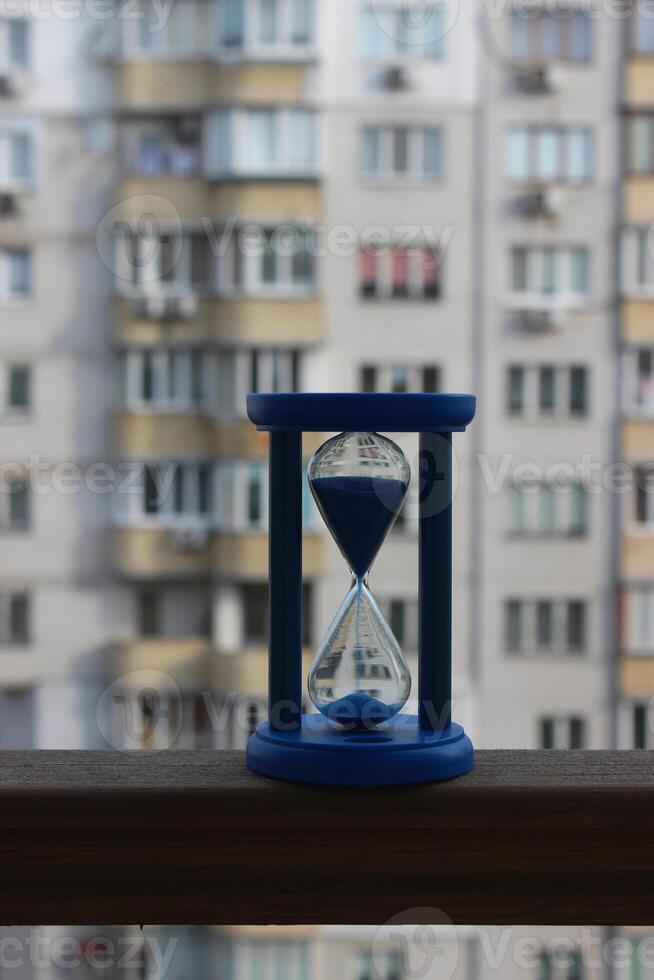 Blue Hourglass With Sand Falling Down On A Wooden Railing Of The Balcony Of A High Rise Building photo