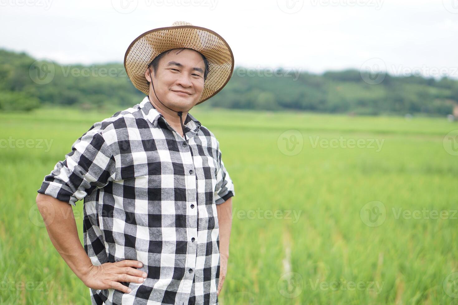 Handsome Asian man farmer is at paddy field, wear hat, plaid shirt, put hands on waist. Confident.Concept, Agriculture occupation. Thai farmer. Working with nature. Organic farming photo