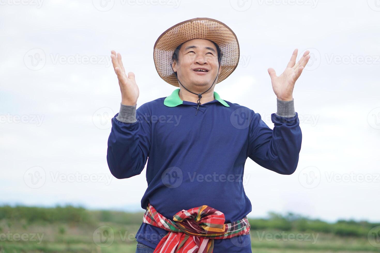 Handsome Asian man farmer stands at agricultural land, wears hat, Thai loincloth on waist, raises hands up with surprised or excited feeling. Concept, agriculture occupation. photo
