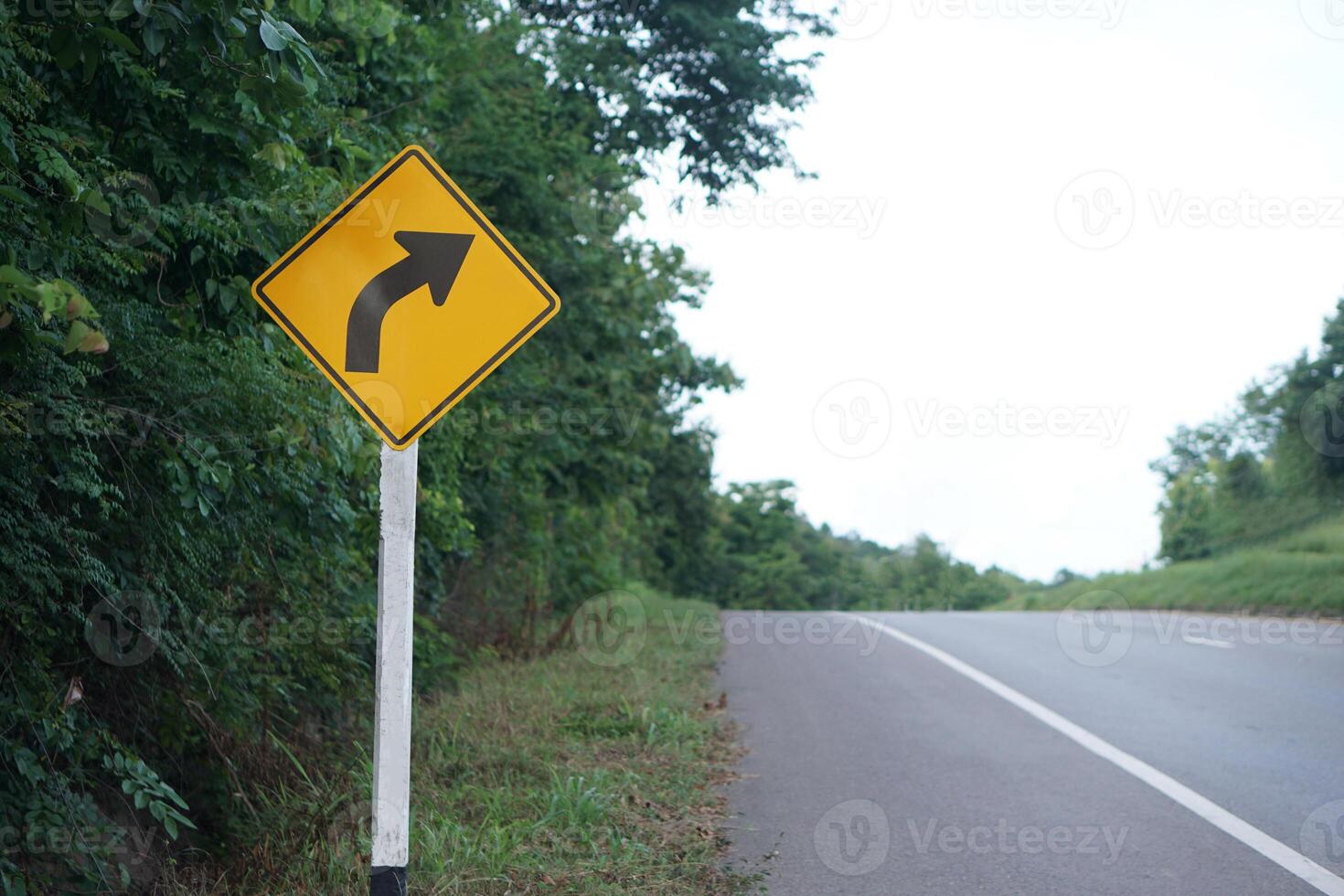 Yellow traffic sign with arrow left curve symbol beside the rural road to warn drivers to know the way ahead has right curve. Concept. Warning traffic sign for transportation. photo