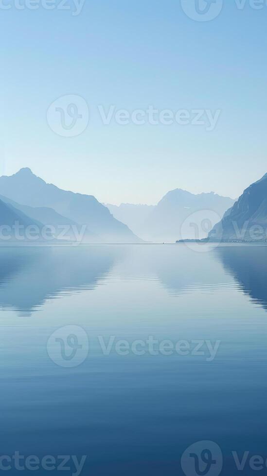 A tranquil lake reflects the silhouette of mountains under a hazy sky photo