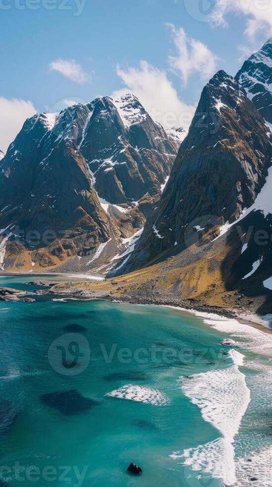 Snowy peaks rise above a turquoise ocean, with whitecaps crashing gently on the shore photo