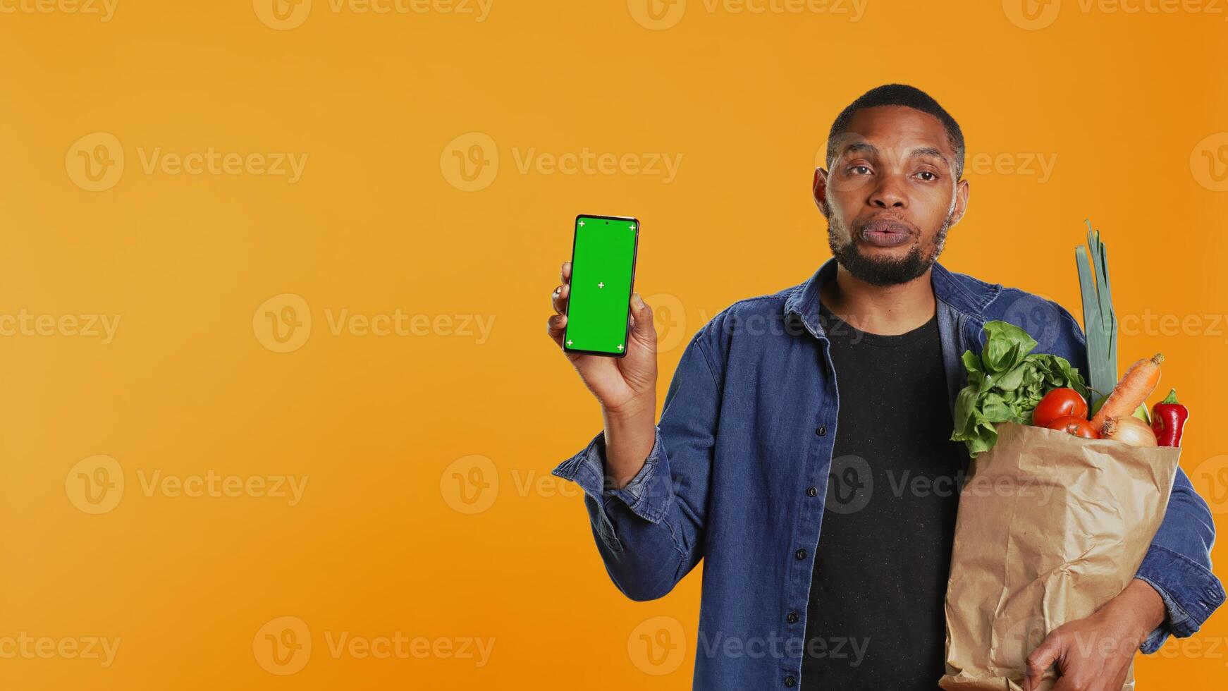 Young adult holding a smartphone with greenscreen layout in studio, carrying bio groceries in a paper bag and advertising healthy eating. Vegan person presenting mockup screen. Camera B. photo