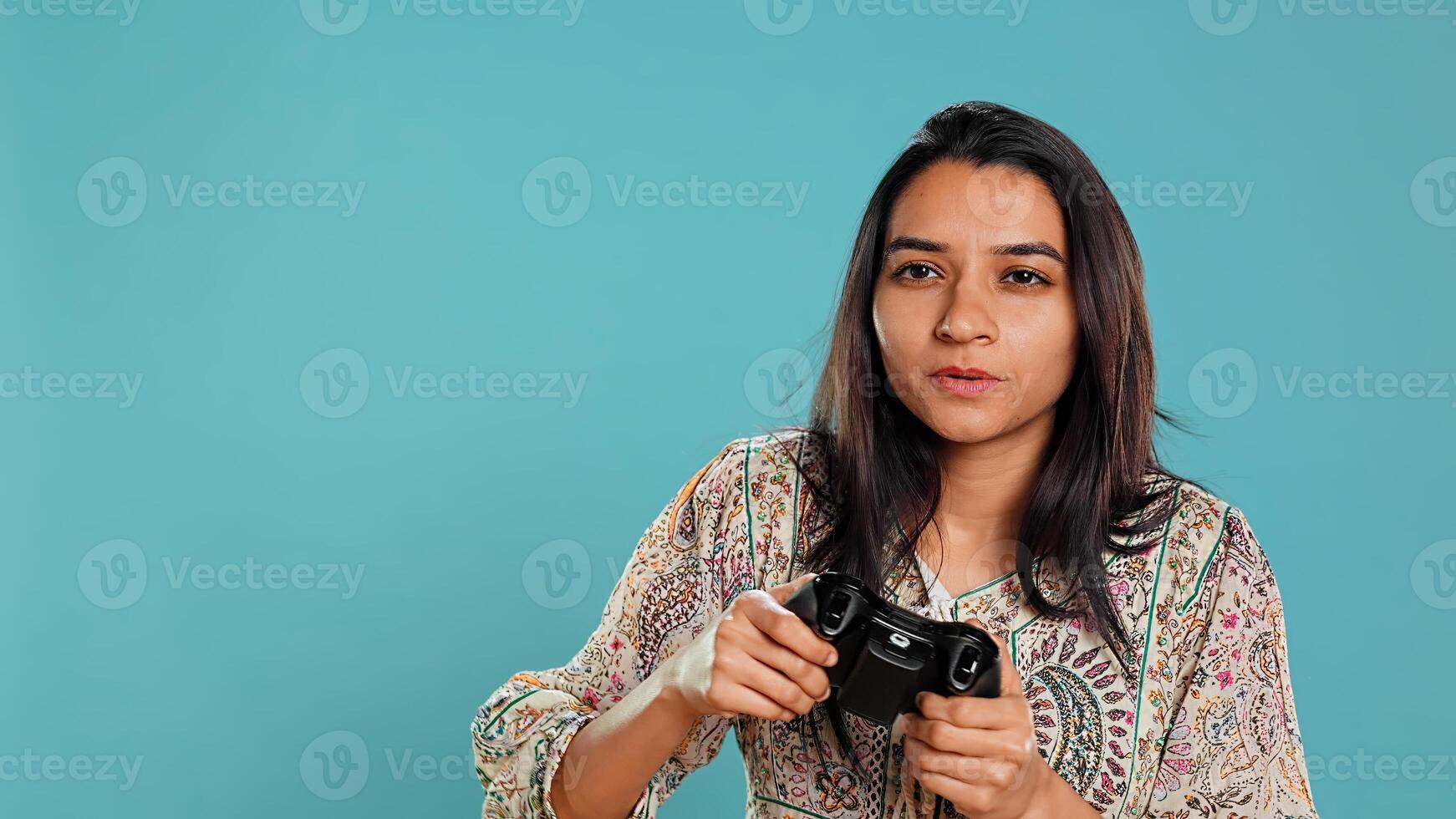 Cheerful indian gamer showing thumbs up sign gesturing while holding controller after winning videogame. Joyful woman enjoying games, doing approving hand gesture, studio background, camera B photo