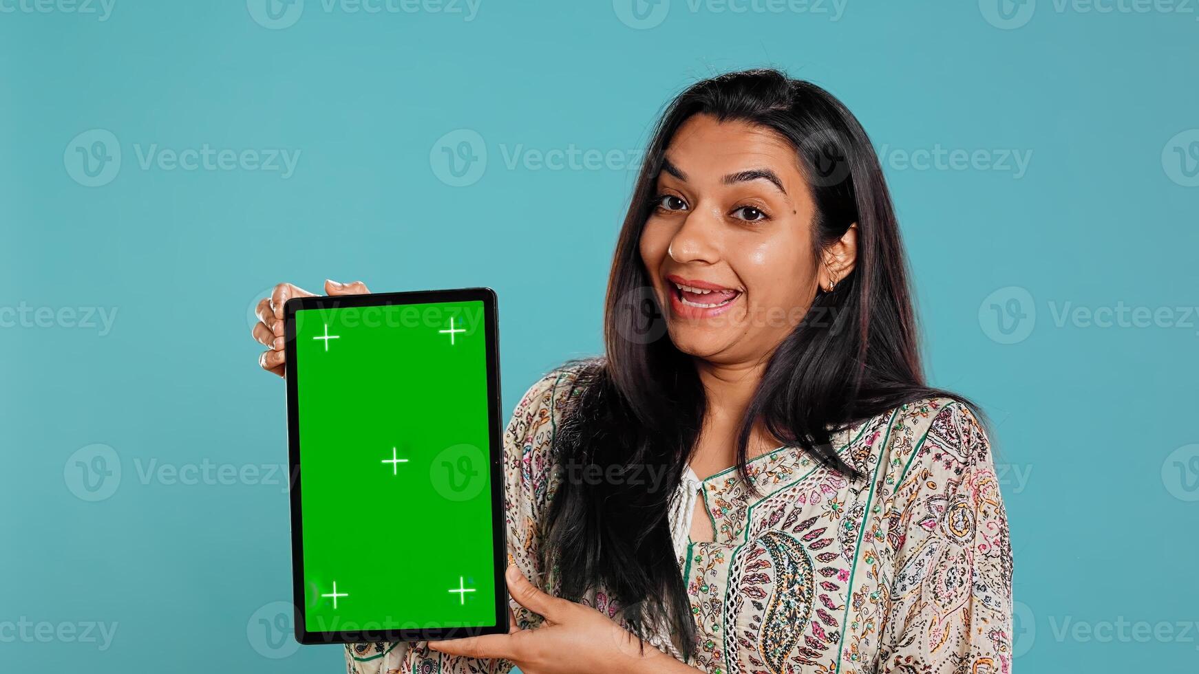 Portrait of indian woman doing influencer marketing using green screen tablet, isolated over studio background. Smiling person holding empty copy space mockup device, camera B photo
