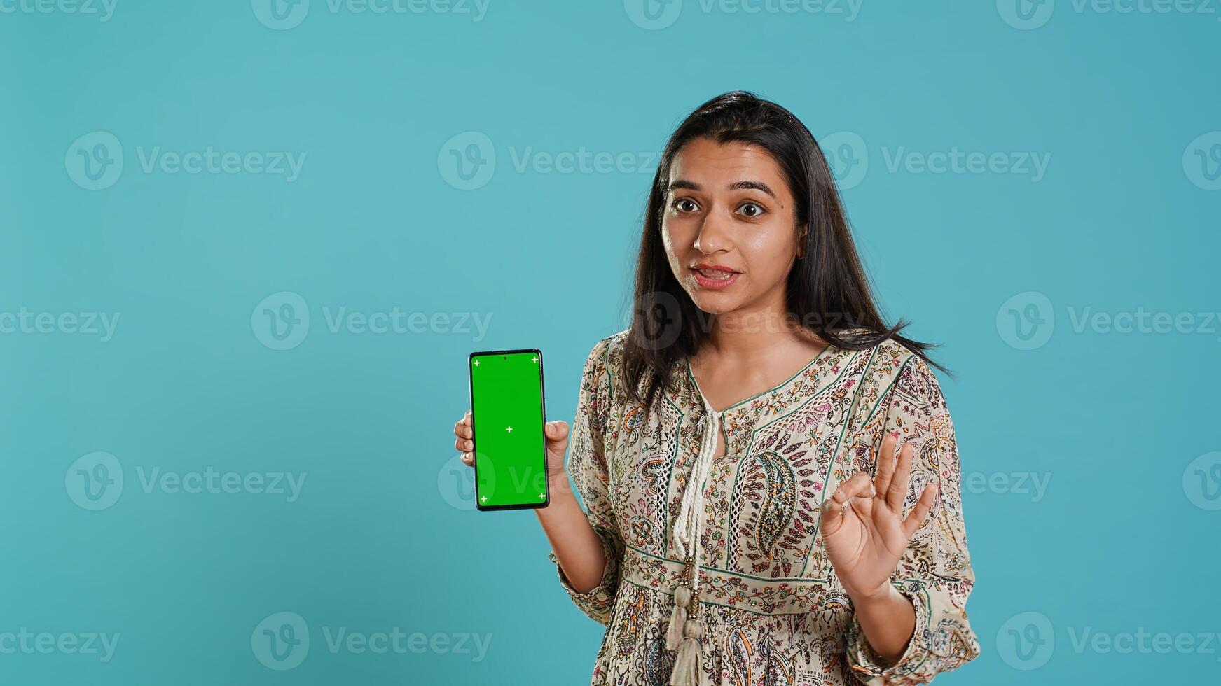Woman presenting green screen mobile phone, isolated over studio background. Cheerful indian person holding copy space chroma key smartphone used for advertising brands, camera A photo