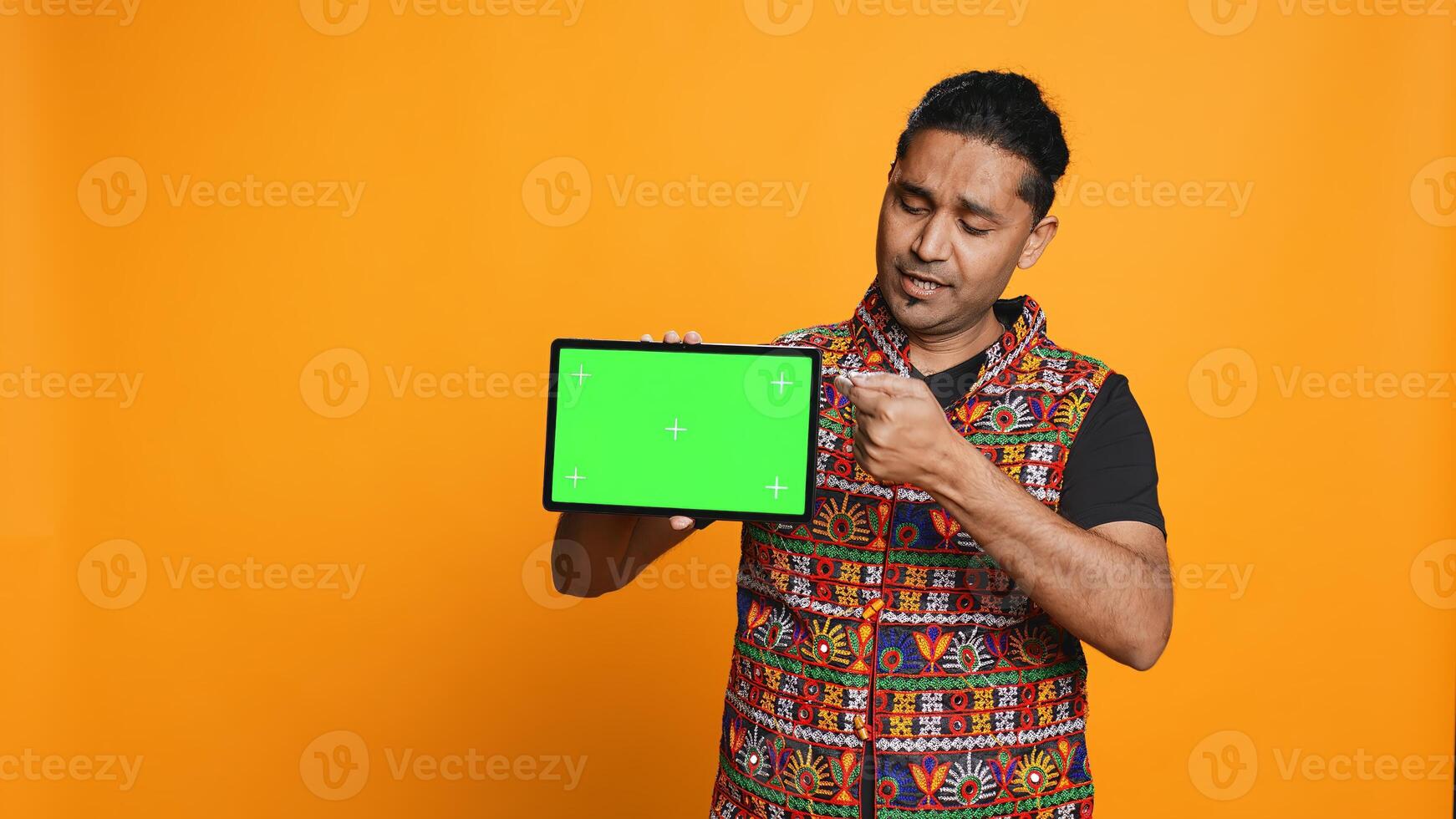Portrait of indian man doing influencer marketing using green screen tablet, isolated over studio background. Smiling person holding empty copy space mockup device, camera B photo