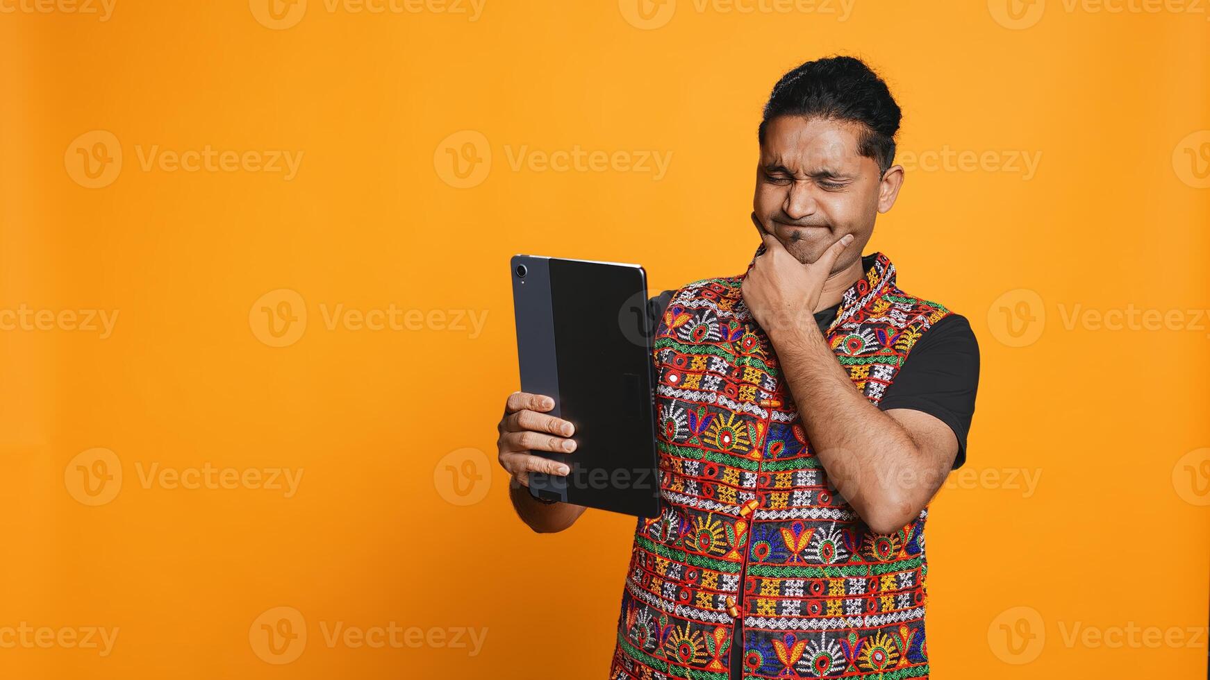 Relaxed man having entertaining conversation with friend over online videocall on device. Carefree indian person discussing with mate on teleconference using tablet, studio backdrop, camera B photo