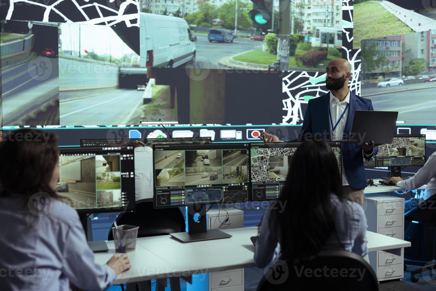 Middle eastern team leader gives instructions to his staff in control center, pointing at satellite map and CCTV footage to guide delivery trucks on their way to the order shipment destination. photo