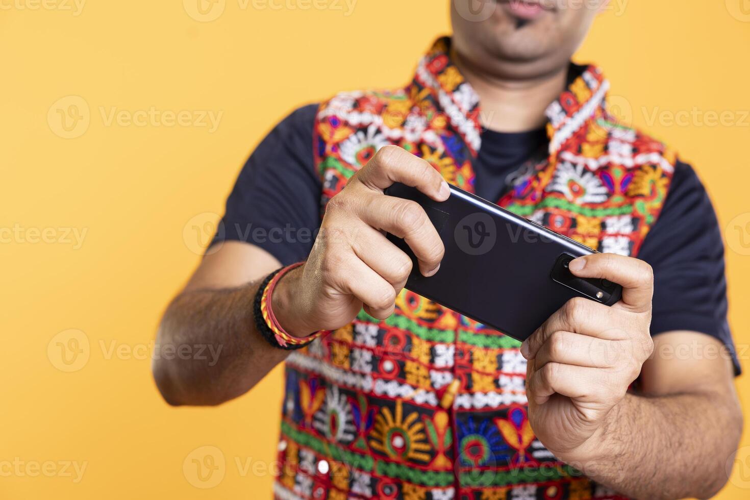 Close up of player playing videogames on smartphone, enjoying relaxation, yellow studio backdrop. Focus on phone used by person in blurry background to do gaming, competing against other players photo