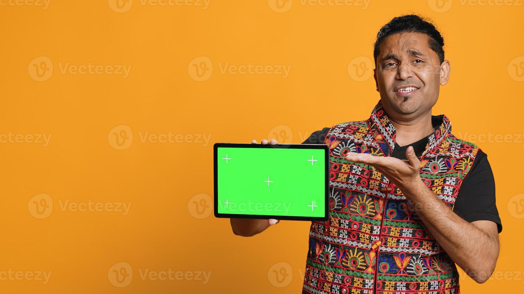 Upset indian man shows thumbs down sign gesturing holding isolated screen tablet, disapproving. Person does rejection hand gesture regarding mockup device, studio background, camera A photo