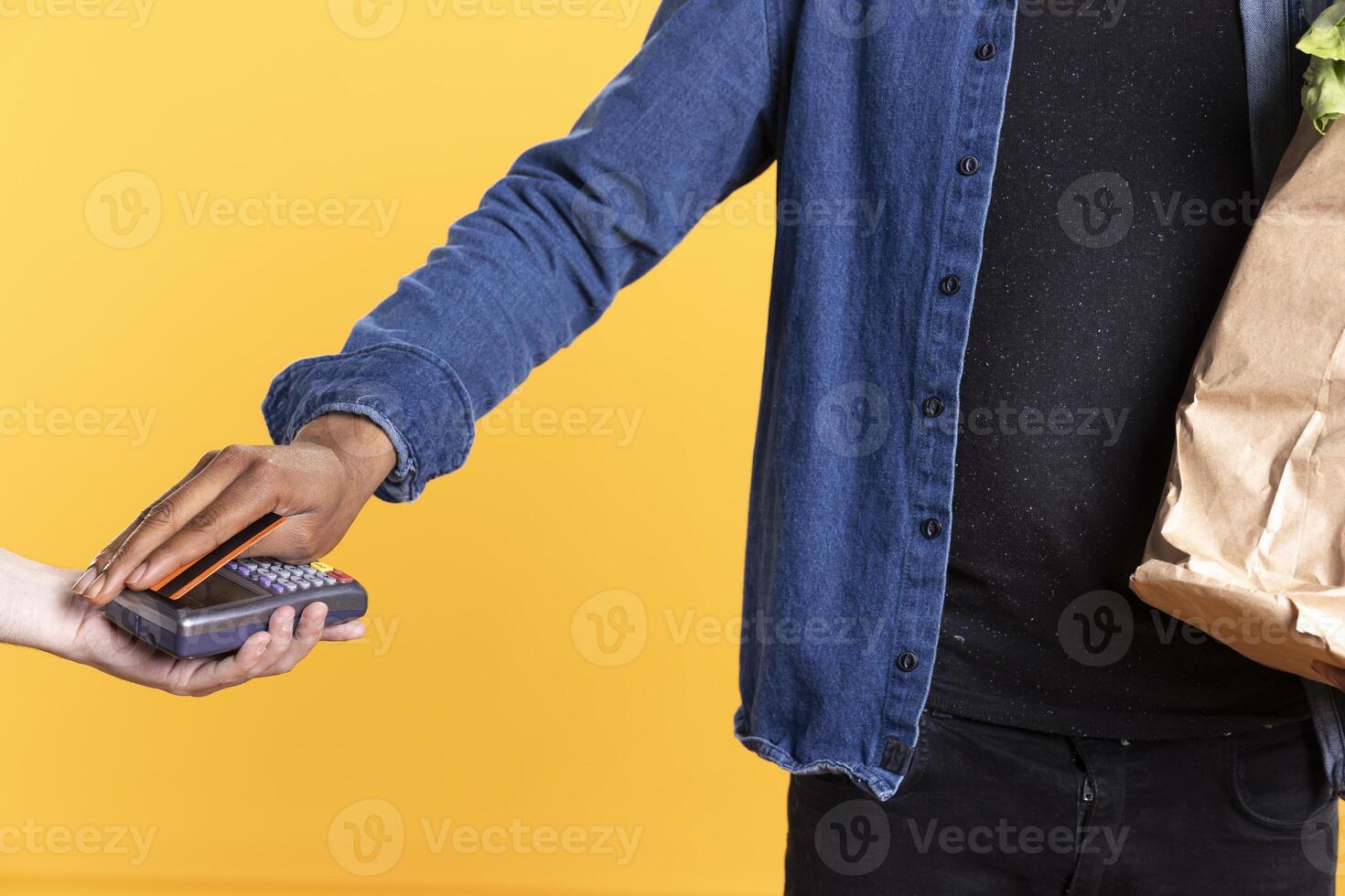 Young person paying with a credit card at the pos terminal on camera, buying fresh ethically sourced produce in a paper bag. Male model using electronic payment during grocery shopping. Close up. photo