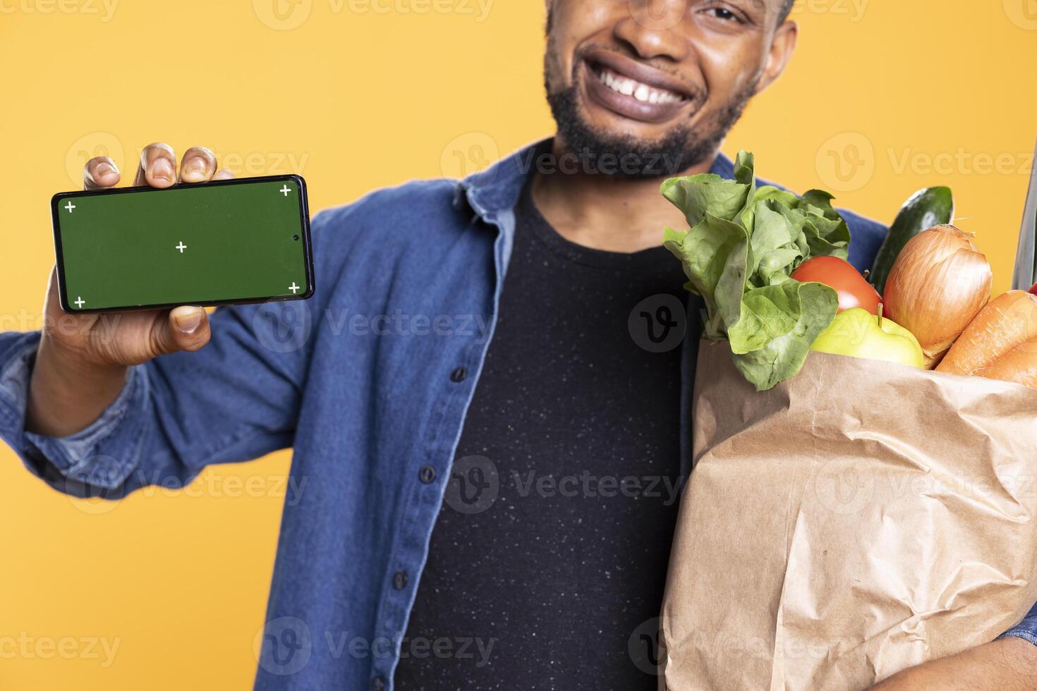 Happy charismatic person showing greenscreen display on camera, carrying a bag of groceries and smiling. Positive vegan guy presenting isolated copyspace template on smartphone. photo
