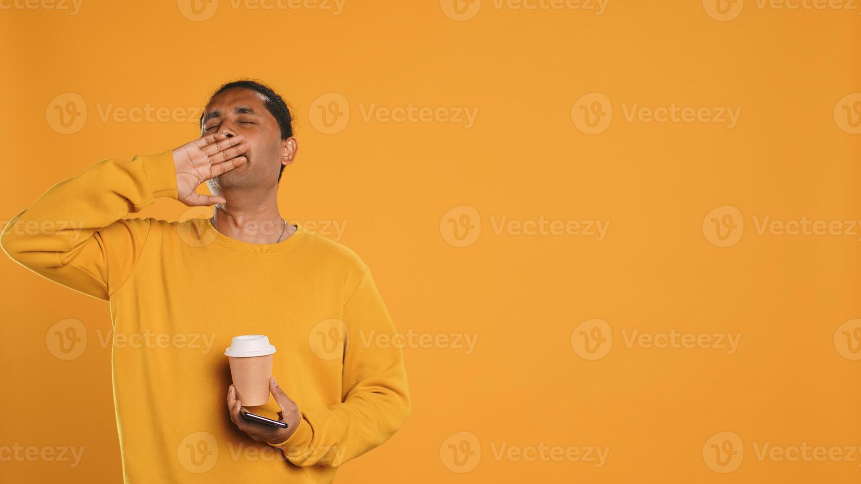 Indian man enjoying coffee from disposable paper cup and send text messages on smartphone. Person drinking hot beverage and chatting with friends on mobile phone, studio background, camera A photo