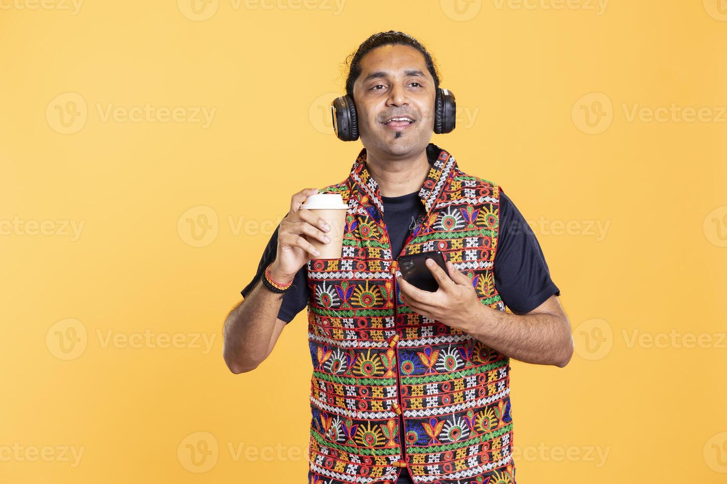 Man sipping coffee and texting friends using mobile phone, enjoying tunes in headphones. Gleeful person enjoying lifestyle, tasting hot beverage, talking with mates using cellphone, studio backdrop photo