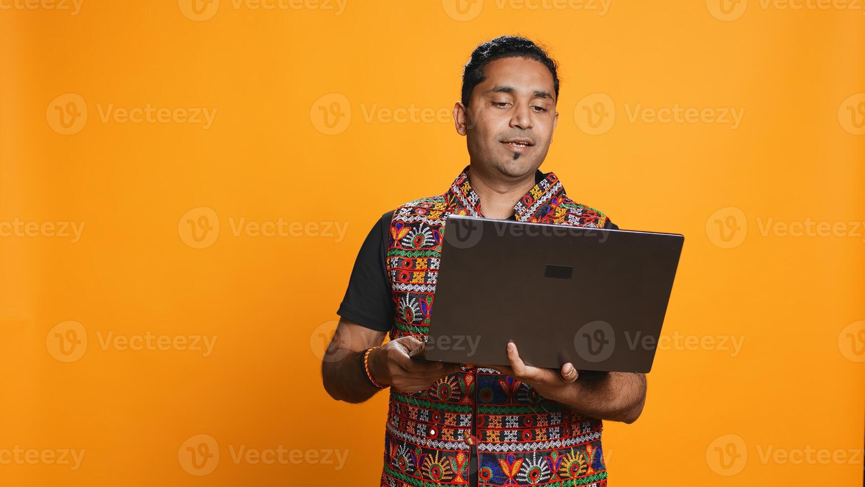 Indian man having entertaining conversation with friends during teleconference meeting using laptop, studio background. Happy person interacts with mates during online videocall on notebook, camera B photo