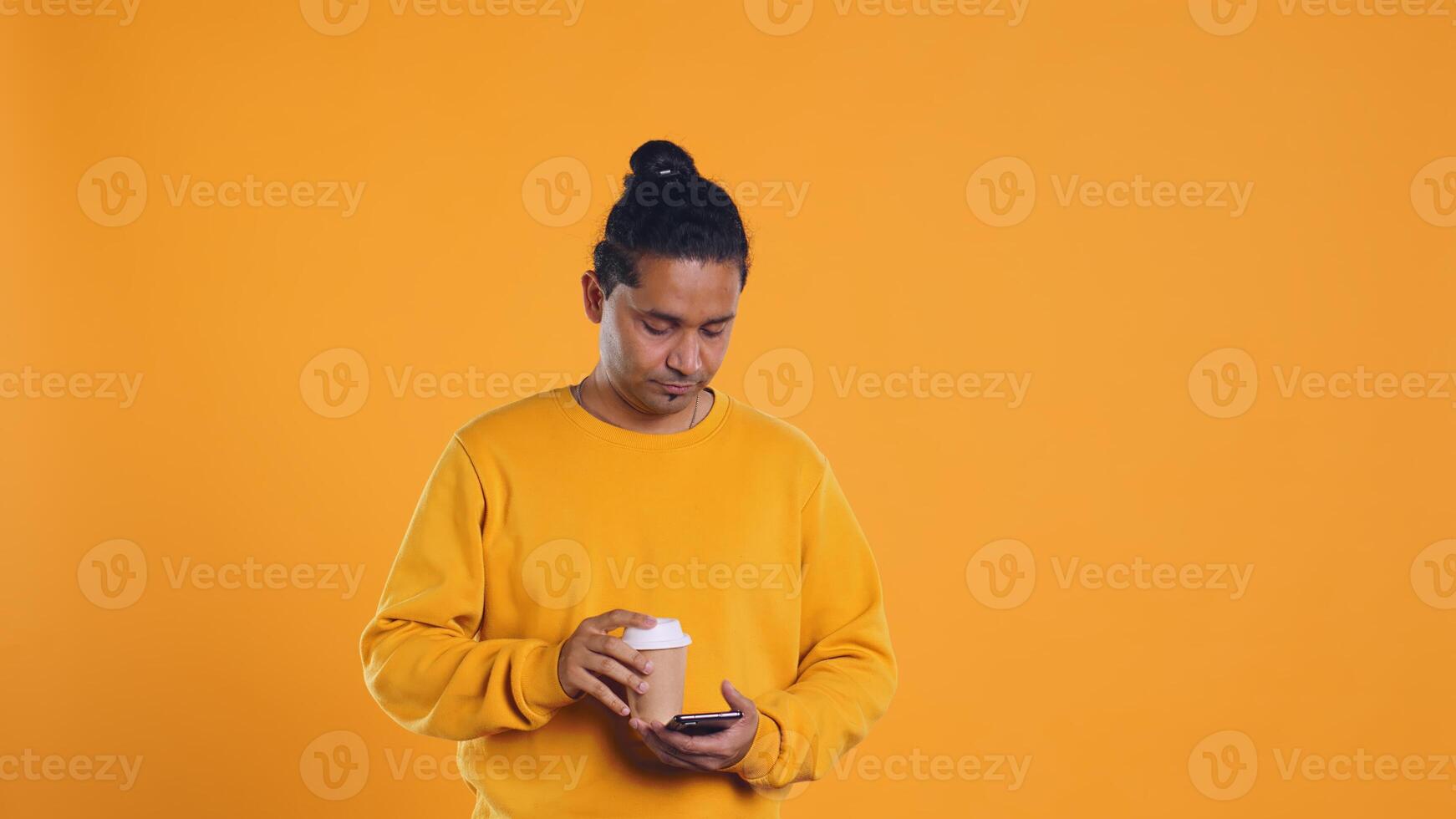 Indian man enjoying coffee from disposable paper cup and send text messages on smartphone. Person drinking hot beverage and chatting with friends on mobile phone, studio background, camera B photo