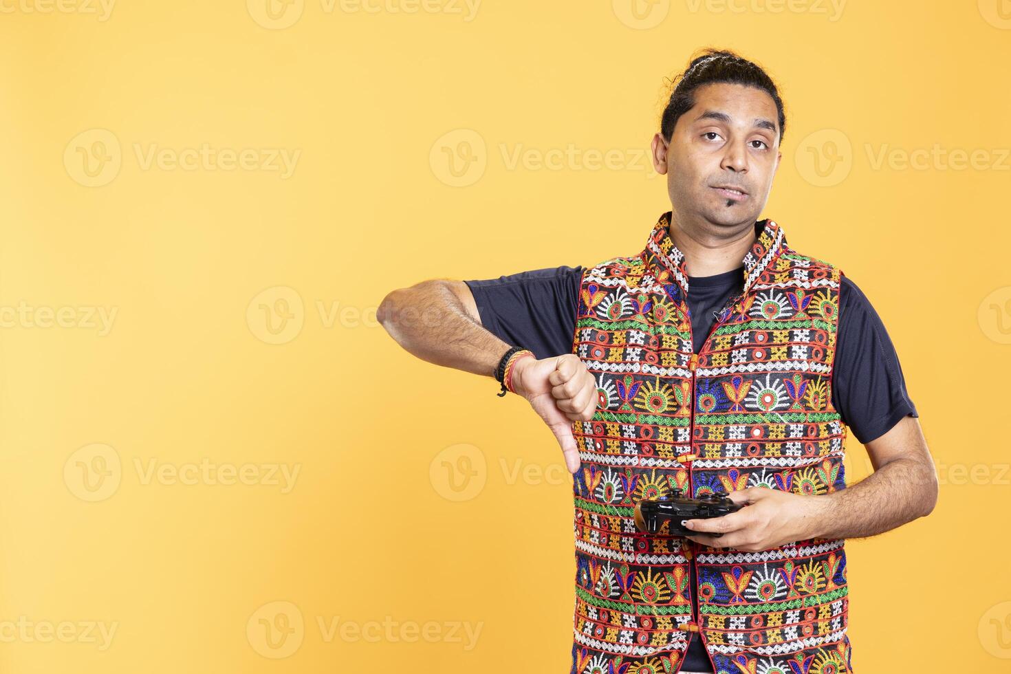 Upset man showing thumbs down sign gesturing holding controller, disapproving of videogames. Displeased person against gaming, doing rejection hand gesture, studio background photo