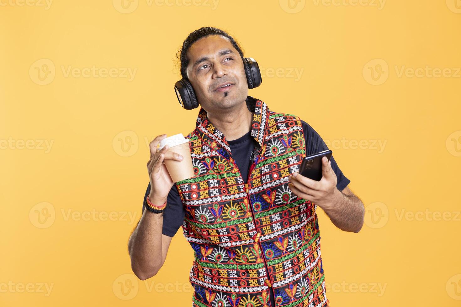 Upbeat man enjoying fresh coffee, texting friends, listening music, doing brainstorming. Happy person using phone while drinking hot beverage, hearing songs, daydreaming, studio backdrop photo