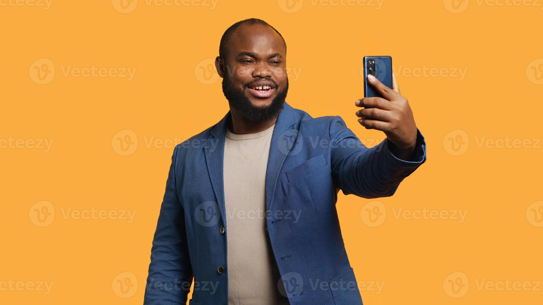 Joyous man using smartphone to take selfies and post them on social media. Happy BIPOC person taking photos using phone selfie camera, isolated over yellow studio background, camera A
