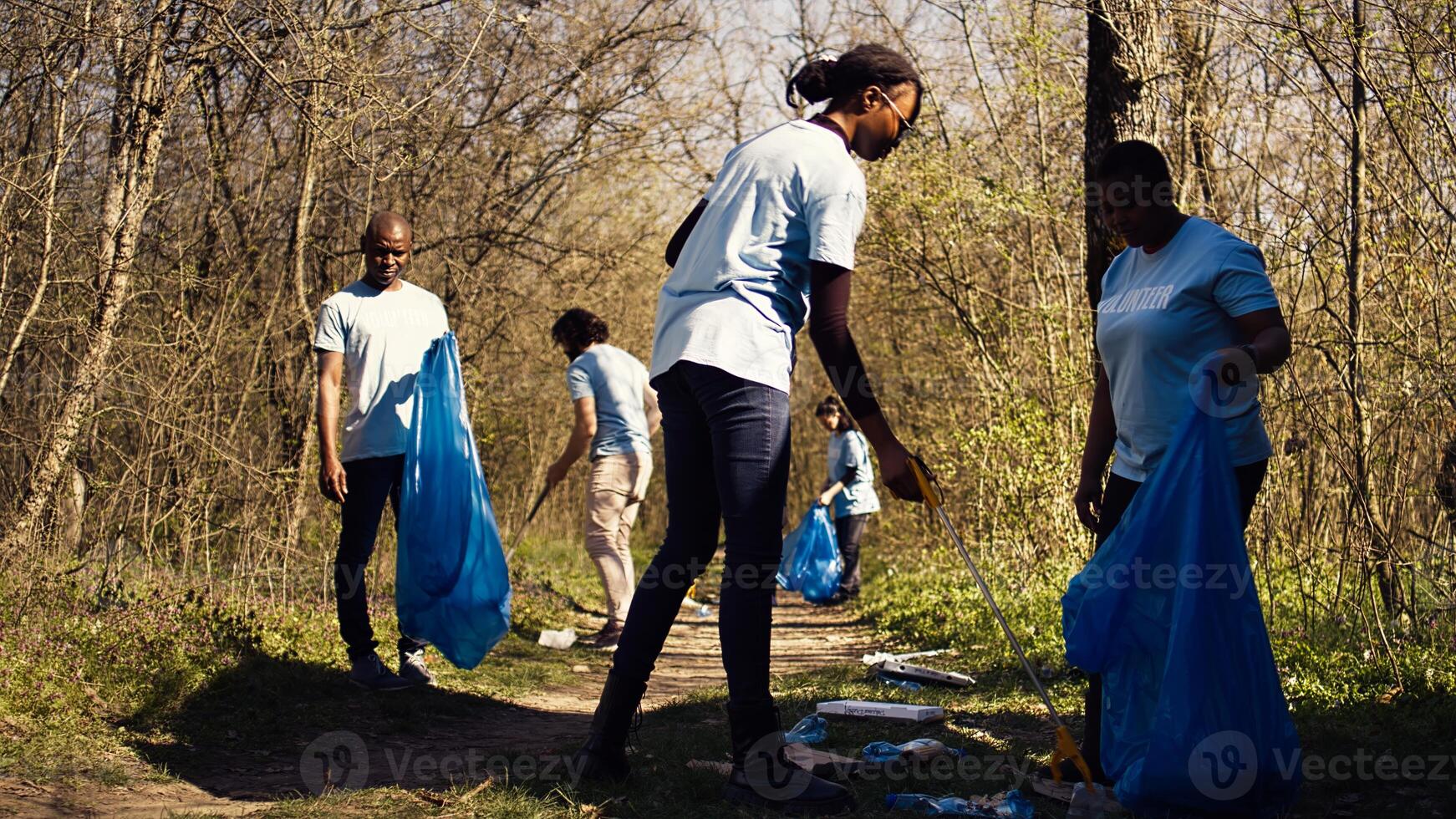 African american team of volunteers picking up trash from the ground, collecting rubbish and recycling in bags. Women cleaning the forest environment from plastic waste, illegal dumping. Camera B. photo