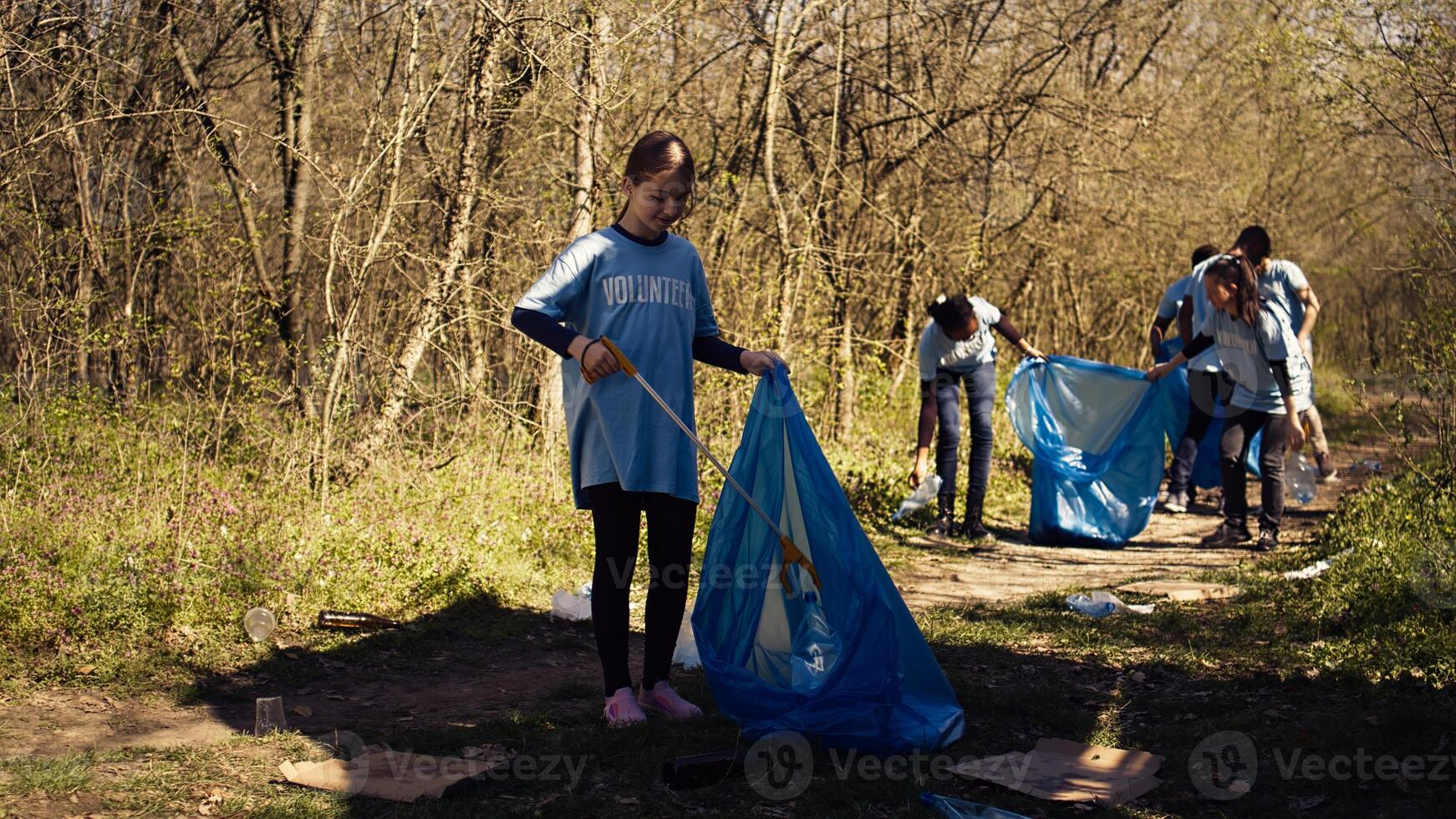 Small girl collecting rubbish around a forest with a claw tool and garbage bags, cleaning up the environment by recycling plastic waste and trash. Child learning to protect ecosystem. Camera B. photo