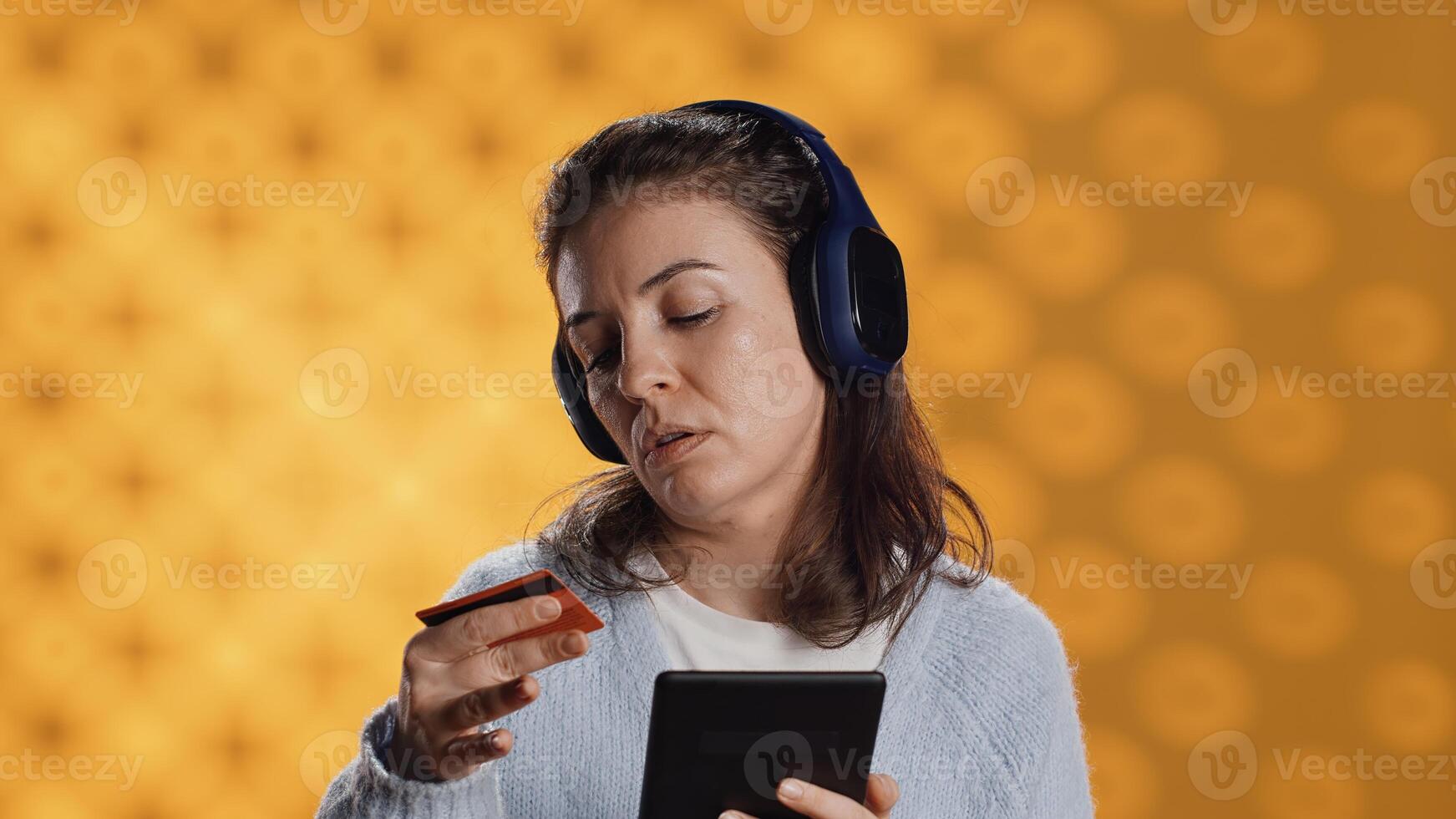 Woman holds ebook reader, listening music and purchasing books online. Person with tablet in hands buying digital novel with credit card, hearing songs in headphones, studio background, camera A photo