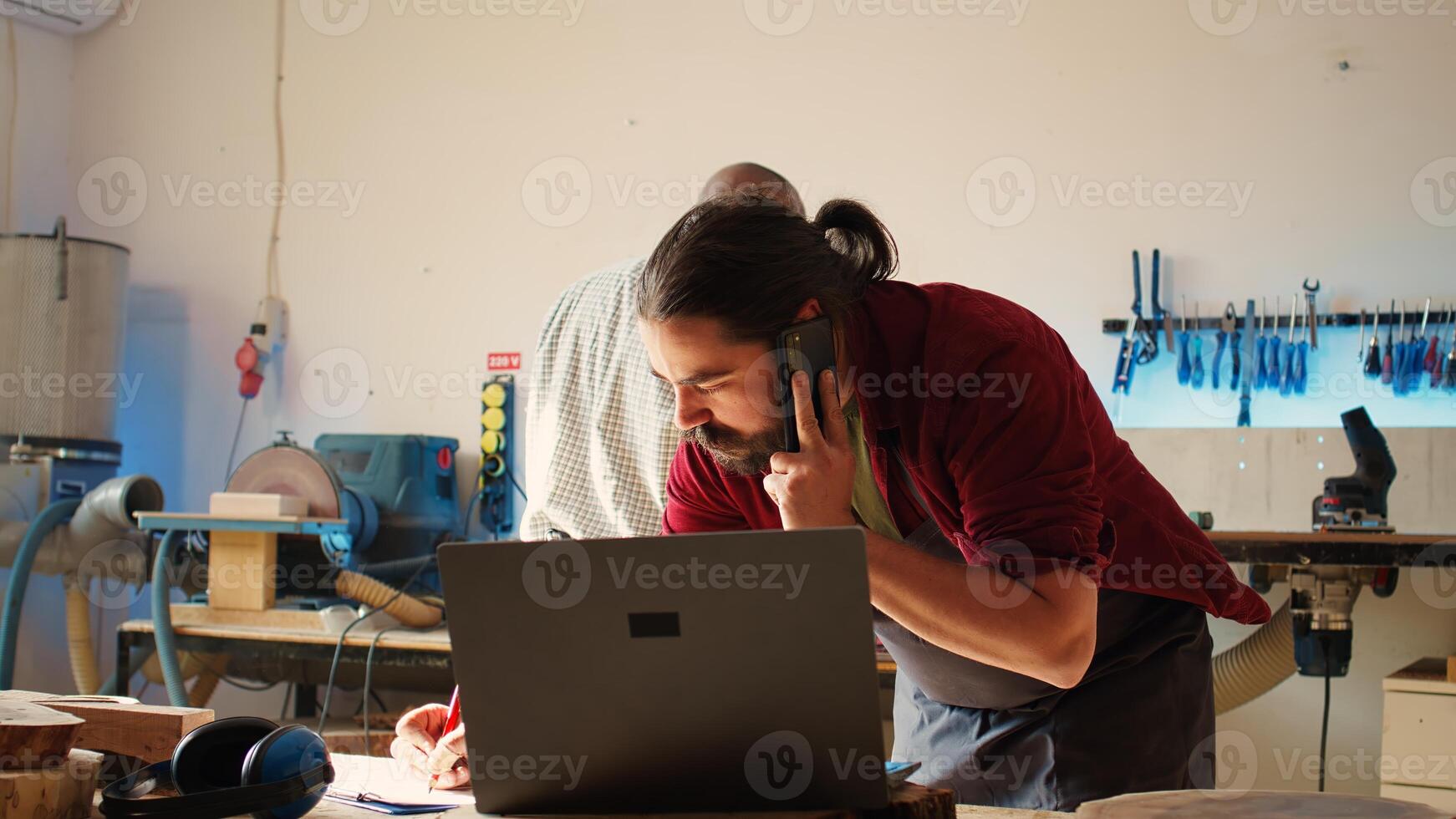 Cabinetmaker receiving telephone call from client commissioning furniture, writing down info. Carpenter in assembly shop drawing schematics, talking on smartphone with customer about order, camera B photo