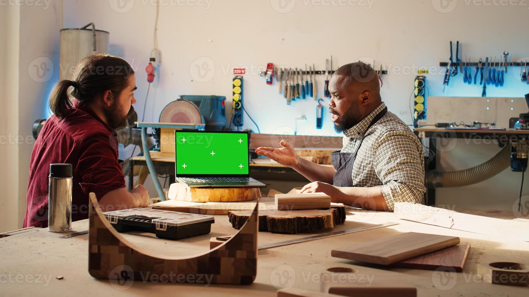 BIPOC artisan and apprentice compare wood art piece with schematic displayed on green screen laptop. Woodworking experts in studio crosschecking wooden object with mockup notebook blueprint, camera B photo