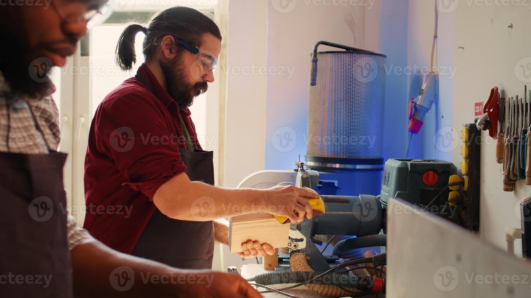 Coworking artisans in studio using sandpaper and wood shaper for sanding wooden surface. Carpenter and african american apprentice wearing safety glasses polishing lumber pieces, camera B photo