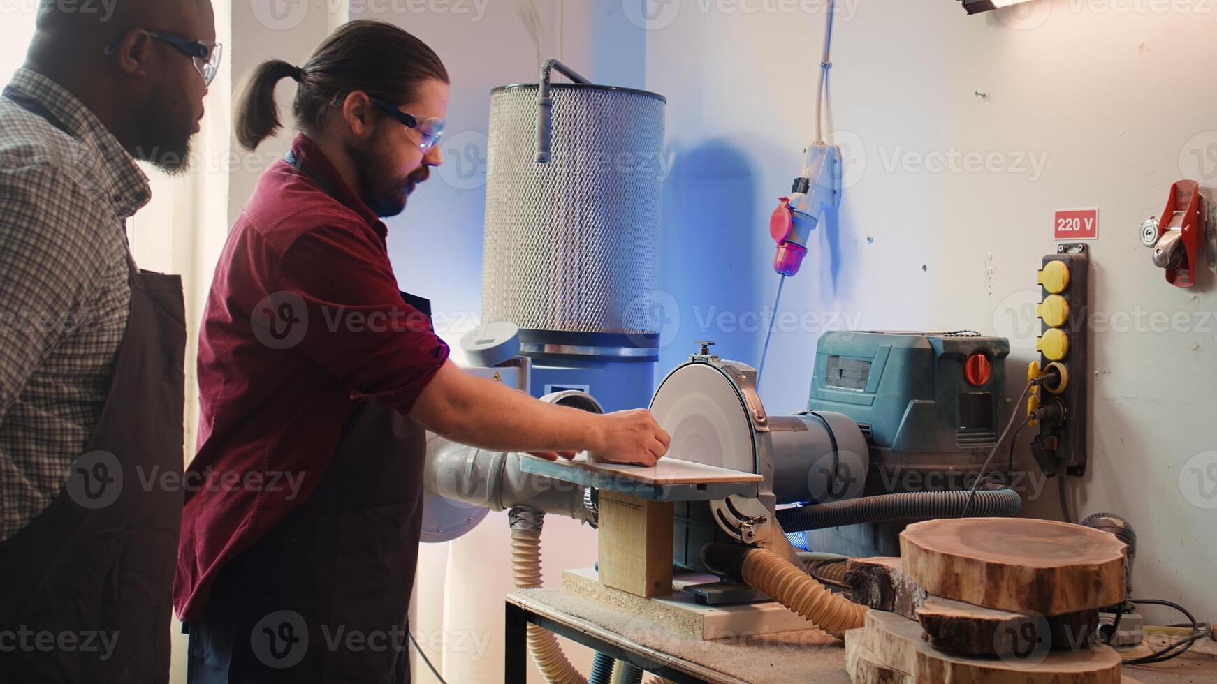 Manufacturer teaching new employee how to shape wooden bowl on disc sander in assembling shop. Apprentice learning from woodworking specialist how to use grinding machine rotating disc, camera B photo