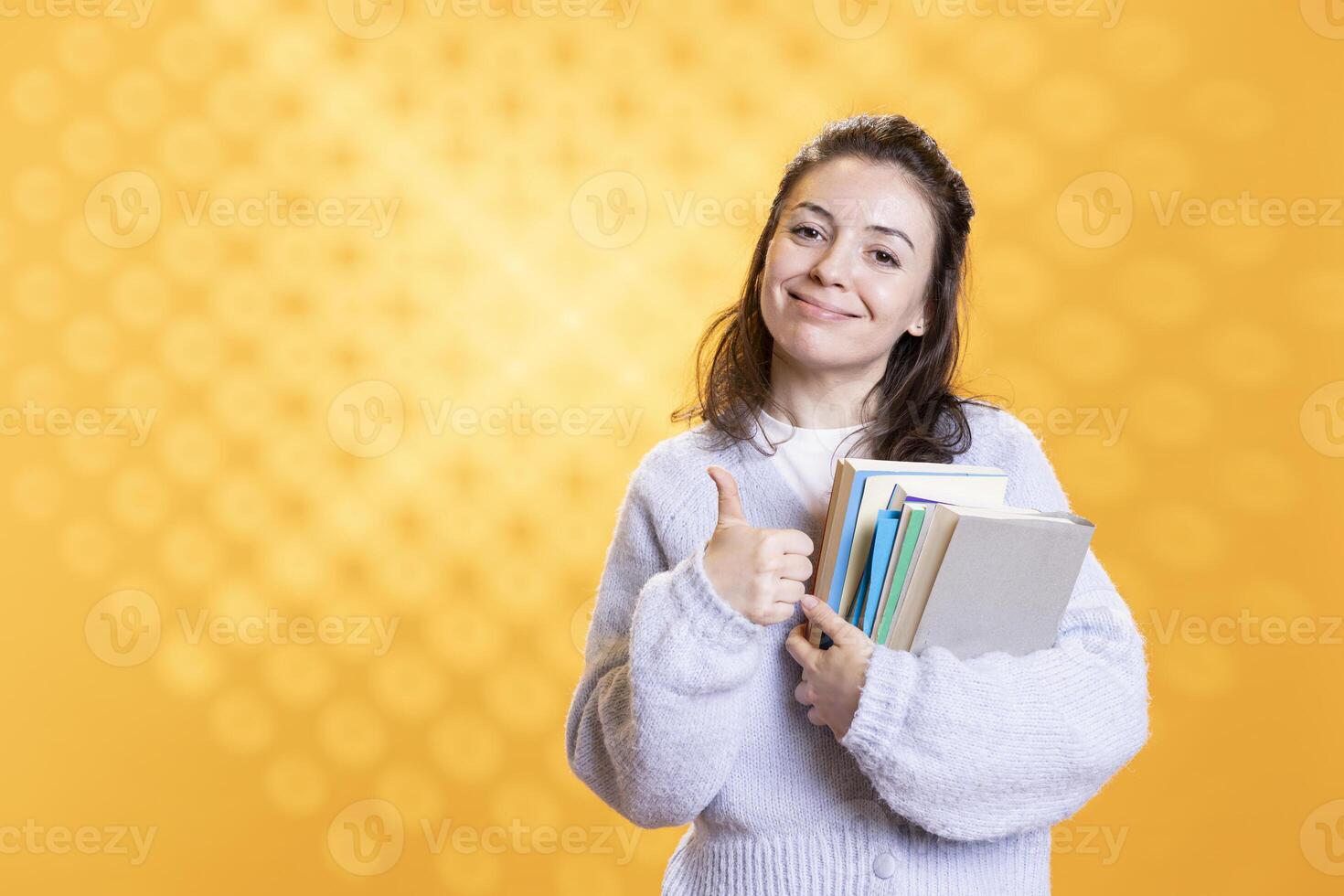 Portrait of happy woman carrying stack of textbooks, gathering information for school exam, studio background, showing thumbs up. Jolly person holding pile of books, doing positive hand gesturing photo