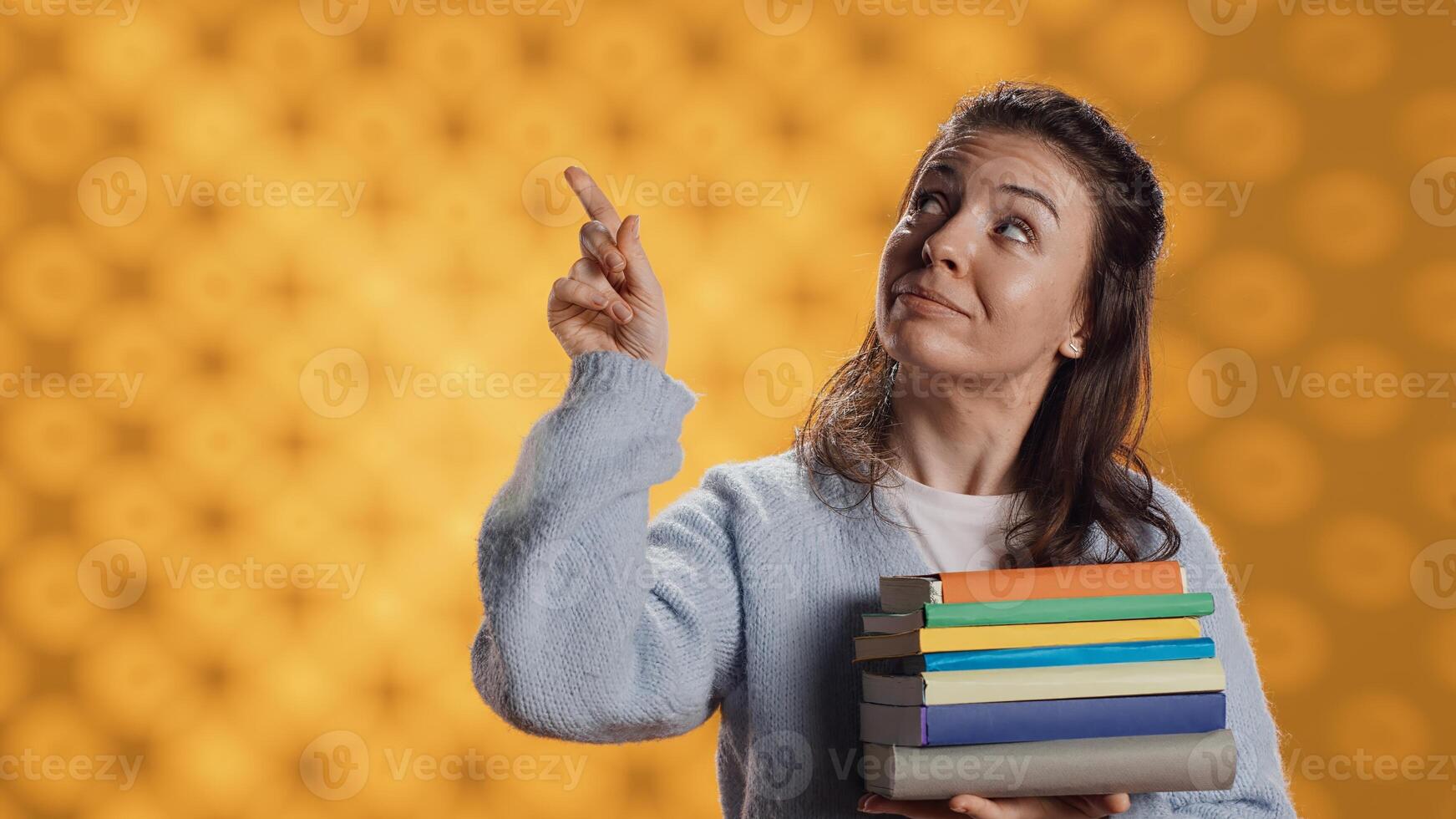 Portrait of smiling woman pointing towards empty copy spaces while holding pile of books. Radiant person with stack of novels in arms promoting reading, showing messages, studio background, camera A photo